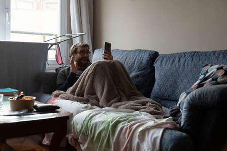 A young man, with a beard and glasses, greets someone on a video call, while resting on the sofa, after eating, covered with a blanket, in the shared apartment in which he lives, Bilbao, Spainの写真素材