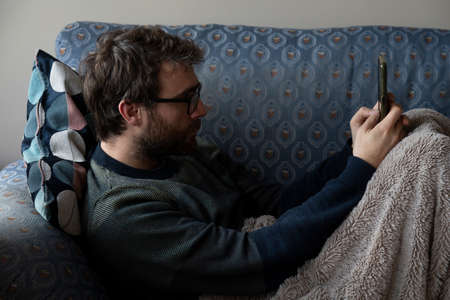 A young man with a beard surfs, chats and reads news on the internet with his smartphone, while he rests on the sofa, covered with a blanket, in the shared apartment where he lives, Bilbao, Spainの写真素材