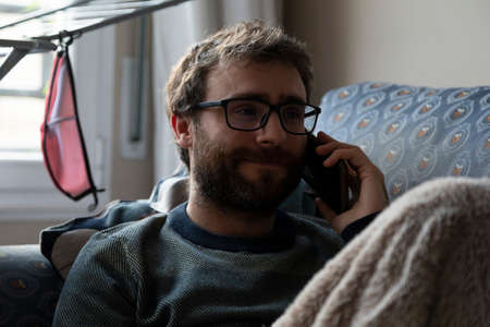A young man, with pasta glasses, bearded, talks on the mobile phone with someone, while resting on the sofa, covered with a blanket, in the shared apartment in which he lives, Bilbao, Spainの写真素材