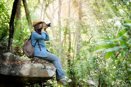 Beautiful Asian girl tourist stands on a rock to photograph nature with a digital camera DSLR in the jungle on a high mountain. Holiday Travel Concept.の写真素材
