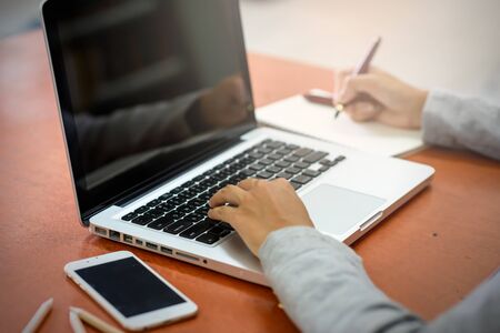 The hands of a young businesswoman using a laptop computer signing a paper on a notebook using a pen. Business and Technology Conceptsの写真素材