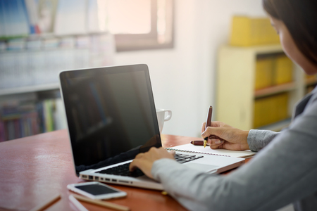 The hands of a young businesswoman using a laptop computer signing a paper on a notebook using a pen. Business and Technology Conceptsの写真素材