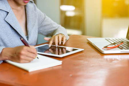 Beautiful Asian businesswoman is using and touching a tablet. At the same time, he wrote the report with a pen on the table to her office. Finance, Banking and Technology Concepts.の写真素材