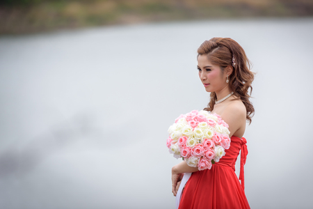 Beautiful woman wear red evening dress hold a bouquet of flowers. over mountains and rivers background.の写真素材
