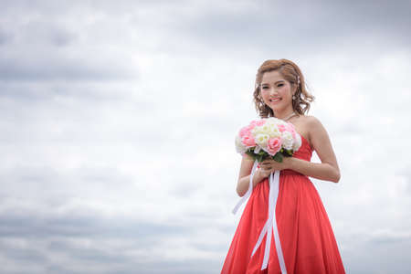 Beautiful woman wear red evening dress hold a bouquet of flowers. over mountains and rivers background.の写真素材