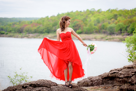 Beautiful woman wear red evening dress hold a bouquet of flowers. over mountains and rivers background.の写真素材