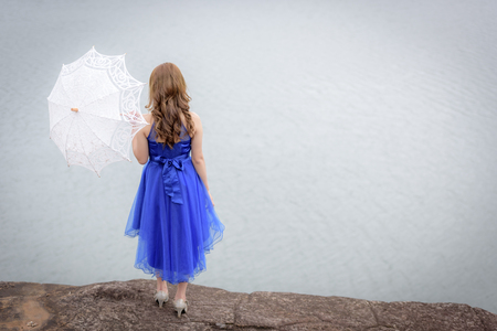 Beautiful woman wear blue evening dress hold white umbrella watching the view on the rocks.の写真素材