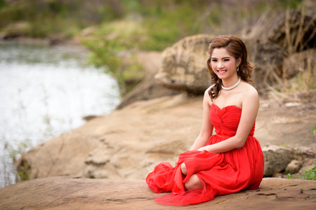 Close up Beautiful woman wear red evening dress happy smile sitting on a rock in the woods.の写真素材