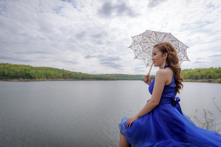 Young woman wearing blue dresses holding a white umbrella while looking at the river and mountains.の写真素材