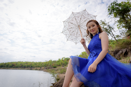 Young woman wearing blue dresses holding a white umbrella while looking at the river and mountains.の写真素材