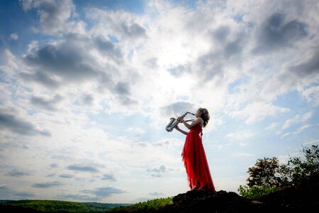 Beautiful woman wear red evening dress sound saxophone stand on the rocks over mountains and rivers background.の写真素材