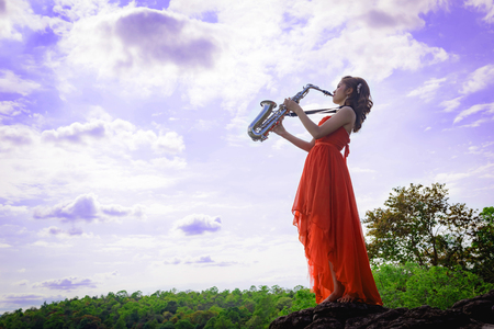 Beautiful woman wear red evening dress sound saxophone stand on the rocks over mountains and rivers background.の写真素材
