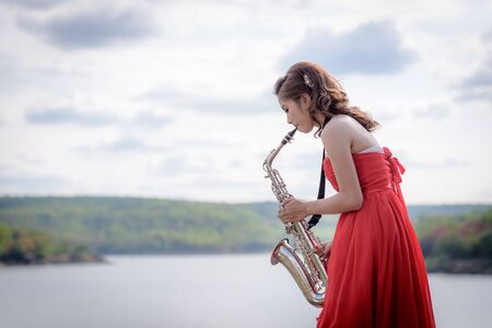 Beautiful woman wear red evening dress sound saxophone stand on the rocks over mountains and rivers background.の写真素材