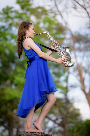 Beautiful woman wear blue evening dress sound saxophone stand on the rocks over mountains and rivers background.の写真素材
