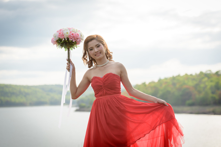 Beautiful woman wear red evening dress hold a bouquet of flowers. over mountains and rivers background.の写真素材