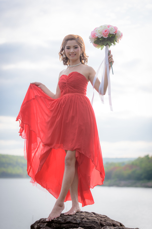 Beautiful woman wear red evening dress hold a bouquet of flowers. over mountains and rivers background.の写真素材