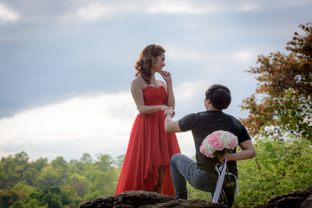 Young man hiding a bouquet of flowers behind him is about to deliver a young woman in red dress.の写真素材
