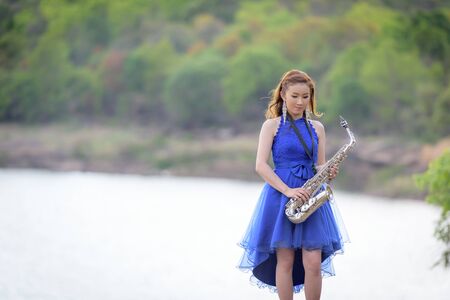 Beautiful woman wear blue evening dress holding saxophone stand on the rocks.の写真素材