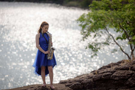 Beautiful woman wear blue evening dress sound saxophone stand on the rocks over mountains and rivers background.の写真素材