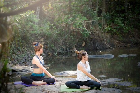 Beautiful mother and daughter are practicing yoga on rocks near the streams, waterfalls. In the rain forest on the mountain. Exercise on vacation Family relationship and nature concept.の写真素材