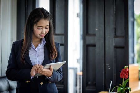 Beautiful Asian businesswoman is using tablet standing near window glass. Within the office. Technology and Business Communication Concept.の写真素材