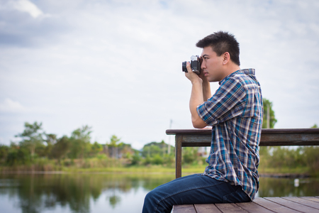 Hipster man sitting on the lake pier with holding vintage camera, Relaxed atmosphere, summer.の写真素材