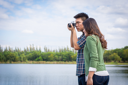 Young men and young women Couple together standing on bench waterfront port in the park. Handle vintage camera.の写真素材