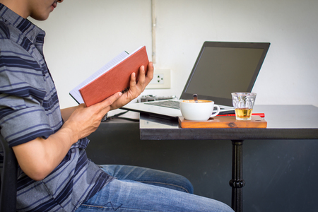 Young man is reading a book with a laptop on the table.の写真素材