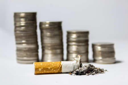 Single cigarette butt with ash and coin stack on white backgroundの写真素材