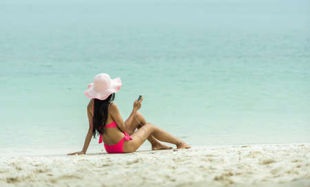 Beach vacation girl listening to music in headphones on a mobile phone during summer travel holiday. Woman relaxing sunbathing on sand sitting wearing sun hat.の写真素材