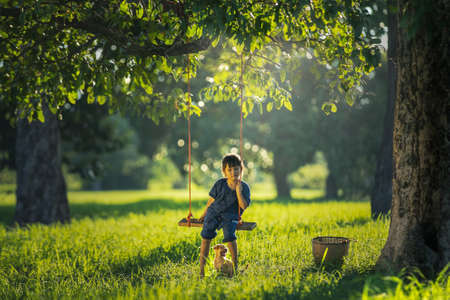 Little Asian girl sitting alone on a swing with her dog, outdoor at countryside of Thailandの写真素材