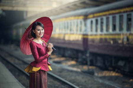 Traveler Asia girl wear traditional dress with umbrella waits for train on railway platform, Bangkok, Thailandの写真素材