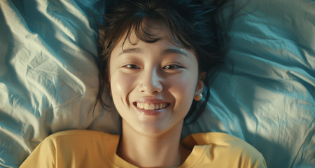 Close-up of happy Asian girl lying in bed on her pillow, smiling and rolling over as if feeling energetic in the morning, waking up early.の素材