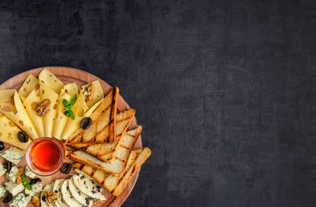 Cheese plate: Parmesan, cheddar, gouda, mozzarella and other with basil on wooden board on dark background with place for text.Honey and Crackersの写真素材