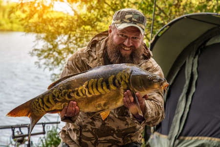 Fishing adventures, carp fishing. Mirror carp (Cyprinus carpio), freshwater fish. Angler with a big carp fishing trophy.Sunrise with a Carp Angler overlooking Lakeの写真素材
