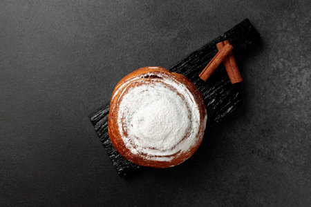 Round, delicious bun with cinnamon and powdered sugar on a black wooden board on a gray background