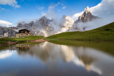 The Pale di San Martino peaks (Italian Dolomites) reflected in the water, with an alpine chalet on background.の写真素材