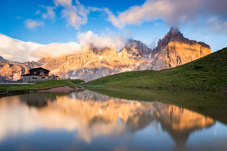 The Pale di San Martino peaks (Italian Dolomites) reflected in the water at sunset, with an alpine chalet on background.の写真素材