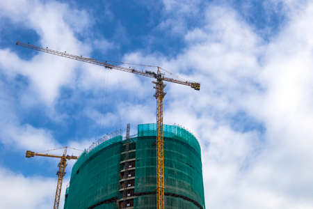 construction of a high-rise building in scaffolding and two yellow construction cranes on the background of the blue cloudy skyの写真素材