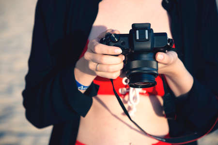 Girl holding modern mirrorless camera in her hands. Hobby and technology concept. Girl stands on the beach and looks at the digital camera.の写真素材