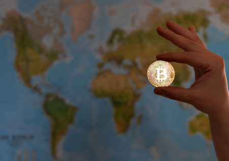 Hand holding gold coin of Bitcoin against map of world. Bitcoin concept. New world currency.の写真素材