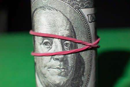one hundred Dollar roll tightened with red rubber band. . Green black background. Franklin looks at...