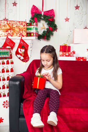 Little girl sitting on the couch holding a red box, Christmas gift, Christmas tree in the backgroundの写真素材
