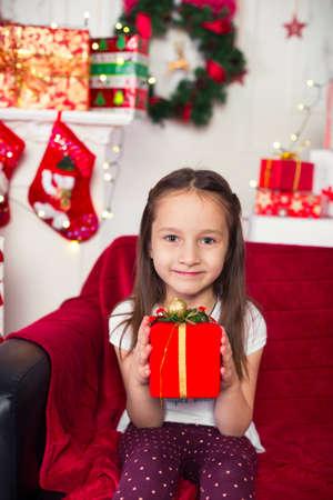 Little girl sitting on the couch holding a red box, Christmas gift, Christmas tree in the backgroundの写真素材