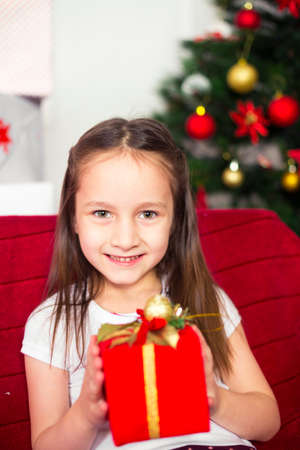 Little girl sitting on the couch holding a red box, Christmas gift, Christmas tree in the backgroundの写真素材