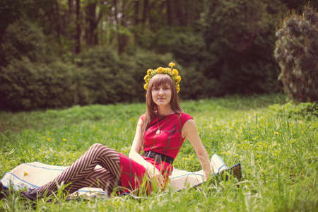 Portrait of beautiful young woman in red dress with dandelion wreath on head sitting in flower field. Outdoor shot.   の写真素材