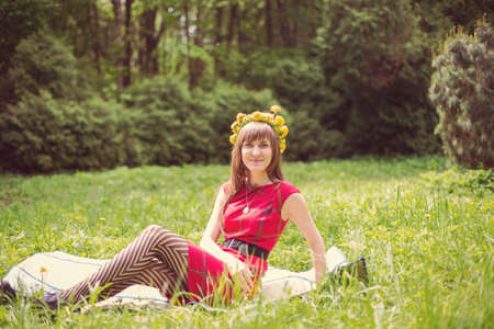 Portrait of beautiful young woman in red dress with dandelion wreath on head sitting in flower field. Outdoor shot.   の写真素材