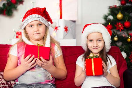 Two girls in Christmas costumes sitting on the sofa with gifts in hand, dressed Christmas tree in the backgroundの写真素材