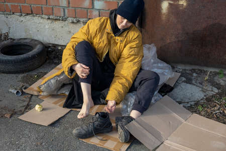 A homeless woman in a yellow old torn jacket and a blue hat sits on a cardboard on the sidewalk and takes off his shoes.の写真素材