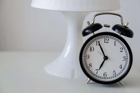 A black alarm clock stands on a white nightstand against a white lamp and white wall. Early rise. Space for text.の写真素材